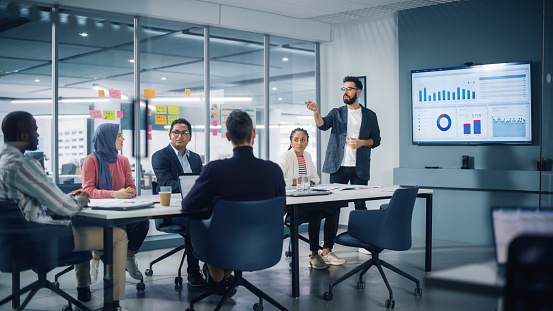 Bureau moderne et diversifié : un homme d'affaires dirige une réunion d'affaires avec les gestionnaires, discute, utilise la télévision de présentation avec statistiques et infographies. Les entrepreneurs numériques travaillent sur un projet de commerce électronique.