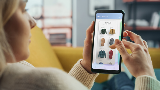 Une belle femme caucasienne utilise un smartphone avec un magasin de vêtements en ligne pour choisir et acheter des vêtements de la nouvelle collection. Femme surfant sur le net et s'allongeant sur le canapé dans le salon de sa maison.