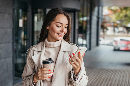 Jeune femme brune ukrainienne souriante tenant un smartphone et une tasse de café et regardant l'écran de l'appareil sur fond d'immeuble de bureaux. copy space