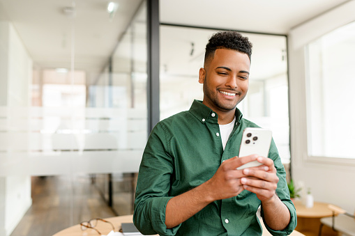 Jeune homme indien, employé, freelance, homme d'affaires, debout, un smartphone à la main, souriant en regardant l'écran, envoyant des textos et des messages en ligne.