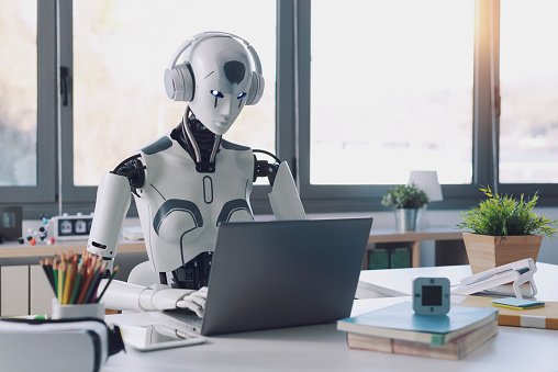 Un robot humanoïde travaille dans un bureau sur un ordinateur portable en écoutant de la musique dans un casque, démontrant l'utilité de l'automatisation dans les tâches répétitives et fastidieuses.