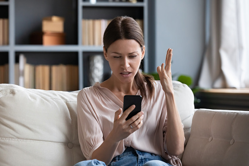 Femme confuse et en colère ayant un problème avec son téléphone, assise sur un canapé à la maison, jeune femme malheureuse regardant l'écran, mécontente d'un smartphone déchargé ou cassé, lisant de mauvaises nouvelles dans un message.
