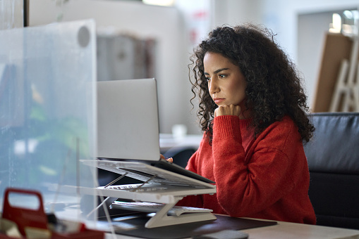 Jeune analyste professionnelle latino-américaine, employée de bureau, assise au bureau, travaillant sur un ordinateur portable, réfléchissant au plan de projet, analysant des données marketing ou financières en ligne, regardant un webinaire d'apprentissage en ligne.