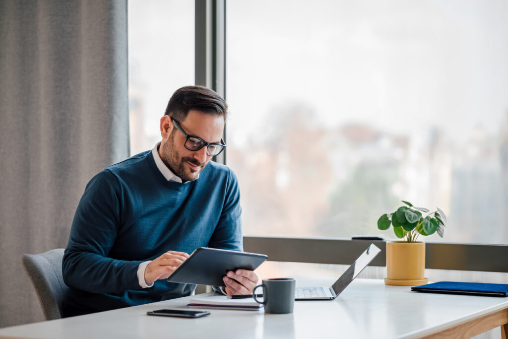 Jeune homme d'affaires utilisant une tablette numérique tout en travaillant sur un ordinateur portable dans un bureau d'affaires. Professionnel masculin avec ordinateur sans fil au bureau. Il est assis près de la fenêtre du bureau.