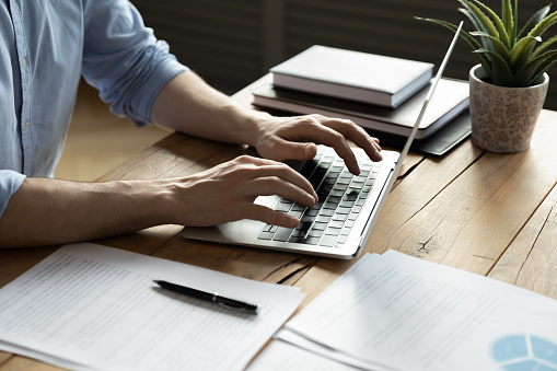 Homme d'affaires en gros plan utilisant un ordinateur portable, tapant sur le clavier, assis à un bureau en bois avec des documents, écrivant des courriels, comptable rédigeant un rapport financier, étudiant occupé à étudier en ligne, recherchant des informations.