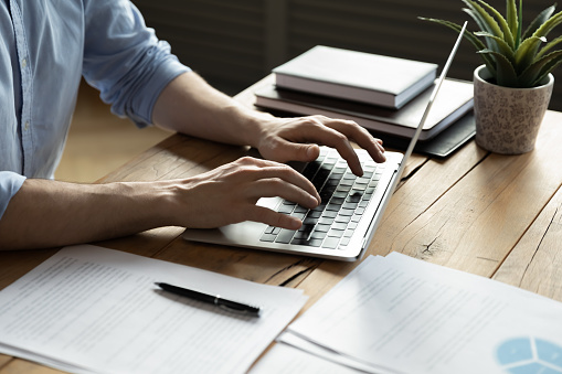 Homme d'affaires en gros plan utilisant un ordinateur portable, tapant sur le clavier, assis à un bureau en bois avec des documents, écrivant des courriels, comptable rédigeant un rapport financier, étudiant occupé à étudier en ligne, recherchant des informations.