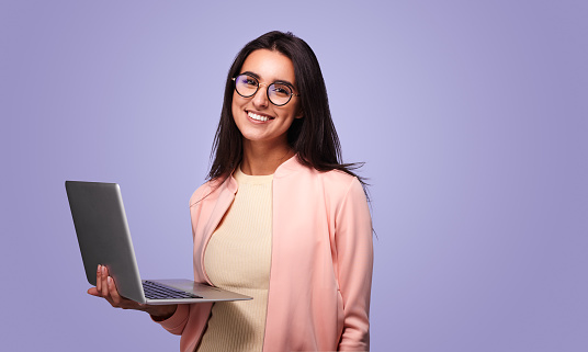 Jeune femme positive, aux cheveux longs et foncés, en tenue branchée et lunettes, souriant et regardant la caméra tout en travaillant à distance sur un ordinateur portable sur fond violet.