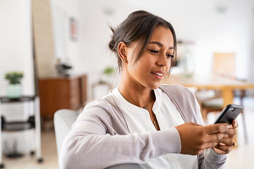 Femme africaine mature assise sur le canapé à la maison utilisant un smartphone. Femme noire se relaxant sur le canapé tout en envoyant des messages avec un téléphone portable à la maison. Femme indienne mature et heureuse tapant sur son téléphone portable dans son salon.