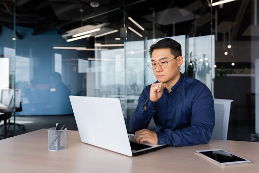 Homme d'affaires asiatique sérieux travaillant dans un bureau moderne, homme mûr en chemise et lunettes utilisant un ordinateur portable au travail, investisseur réfléchissant à une décision complexe.
