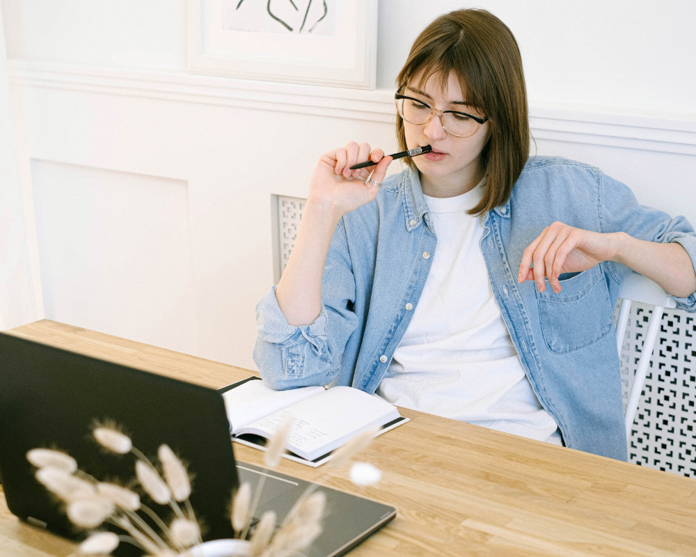 a woman sitting at a table with a laptop and pen in her mouth